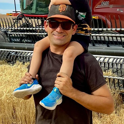Man carries child on shoulders beside wheat harvester